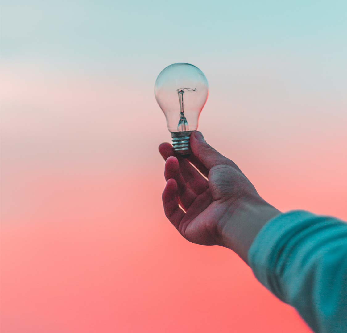 Inspires: a human hand holding a lightbulb in front of a beautiful backdrop of a sunset sky filled with pink and blue colours.